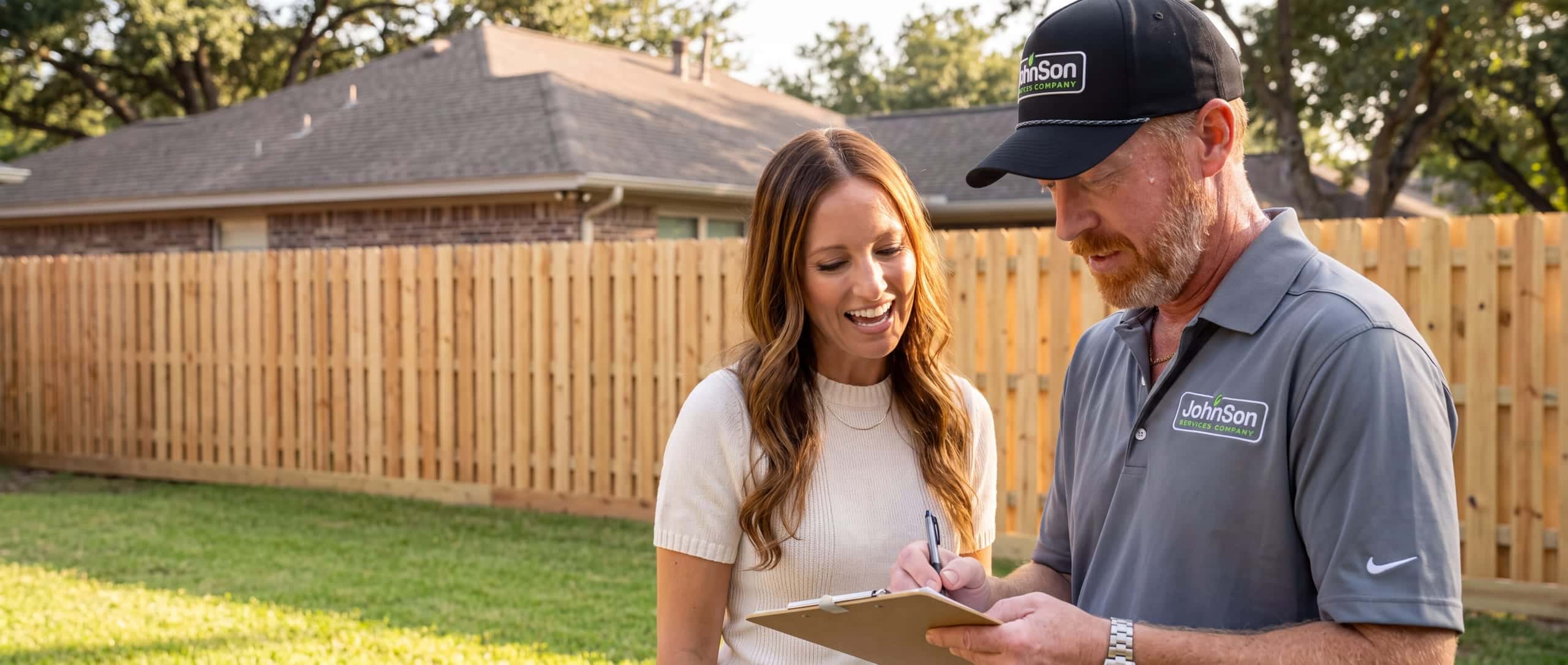 A Johnson estimator writing on a clipboard beside a homeowner in front of a newly built wood fence at an established Texas home